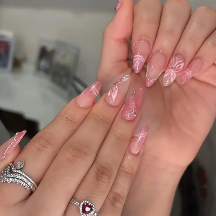 An intricate manicure on long almond nails, with pink cat eye french tips, white abstract floral designs, and rhinestone embellishments.
