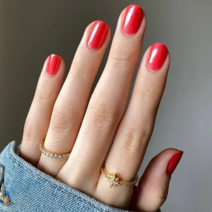 A hand with a shimmering poppy-red manicure on short, natural nails, resting on a denim sleeve.