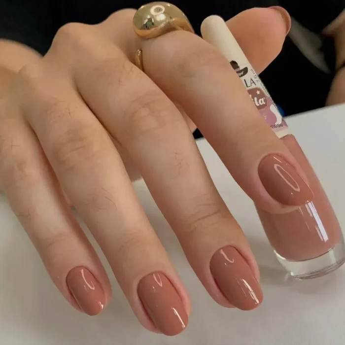 A hand holding a nail polish bottle, showing off a glossy, warm rosy-brown manicure on medium-length nails.