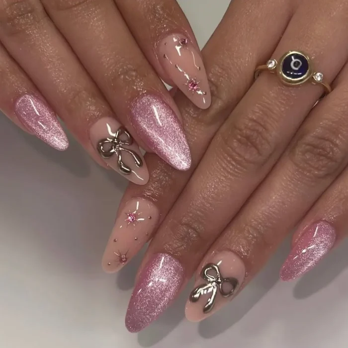 A trendy pink cat eye manicure on long almond nails, featuring 3D silver bow charms and pink rhinestone details.