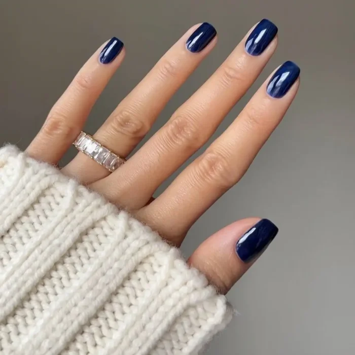 A hand resting on a chunky white knit sweater, showcasing a glossy midnight blue manicure on short, square-shaped nails.