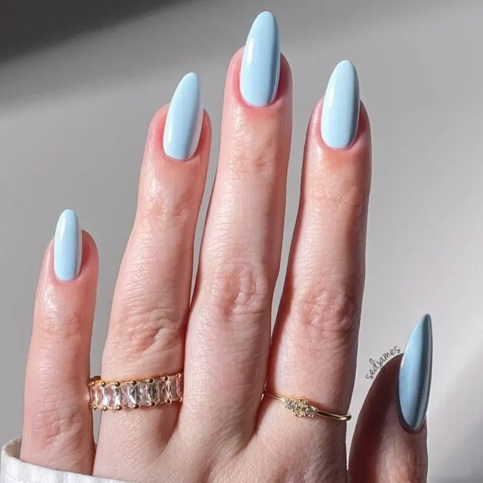 A close-up of a hand with long, almond-shaped nails wearing a bright sky-blue manicure and delicate gold rings.