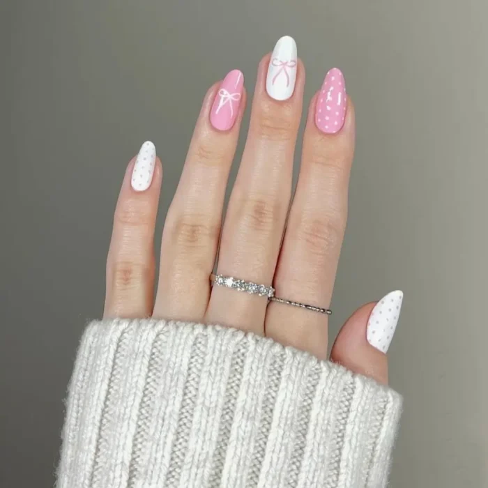A cute white and pink nail design on almond nails featuring polka dots and bow art.