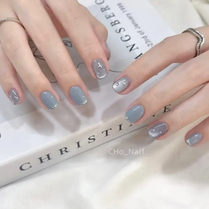 Short, oval nails painted in a soft blue-grey gel, with some nails featuring a subtle shimmer and delicate silver star and moon charms.