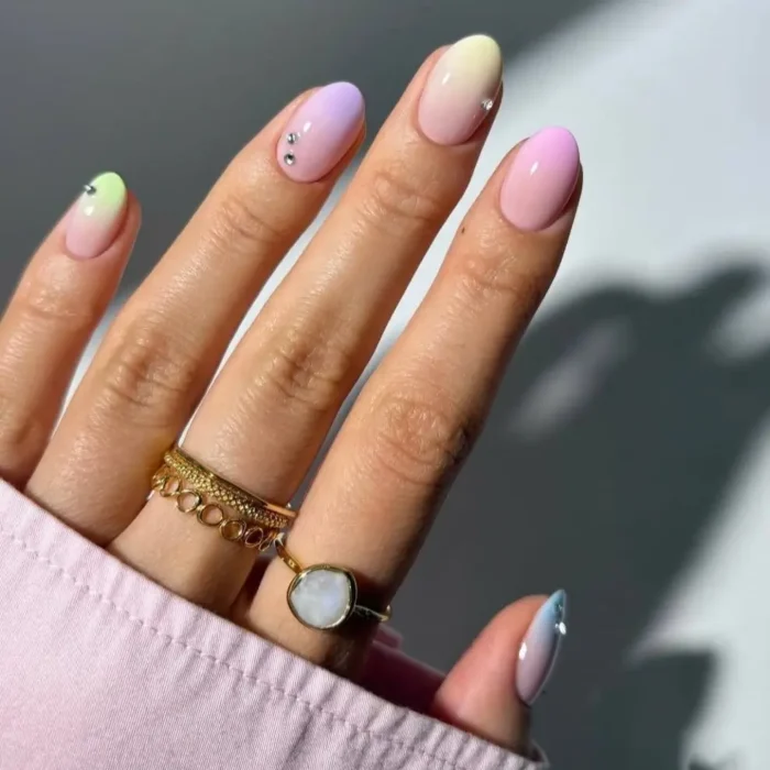 Short, oval-shaped nails with a pastel ombre gel polish, transitioning from yellow to lilac to pink, accented with small rhinestones.