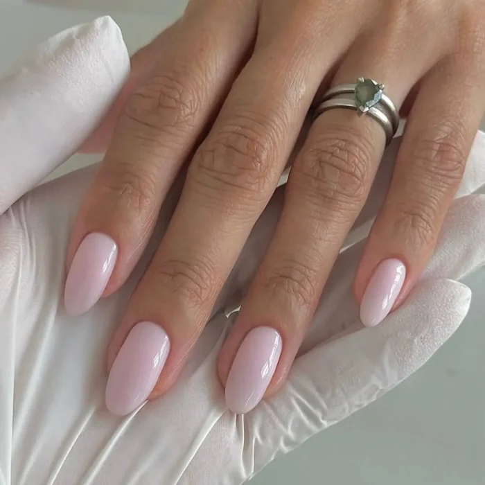 A close-up of a hand with a sheer, milky-pink manicure on perfectly shaped oval nails, resting on a gloved hand.