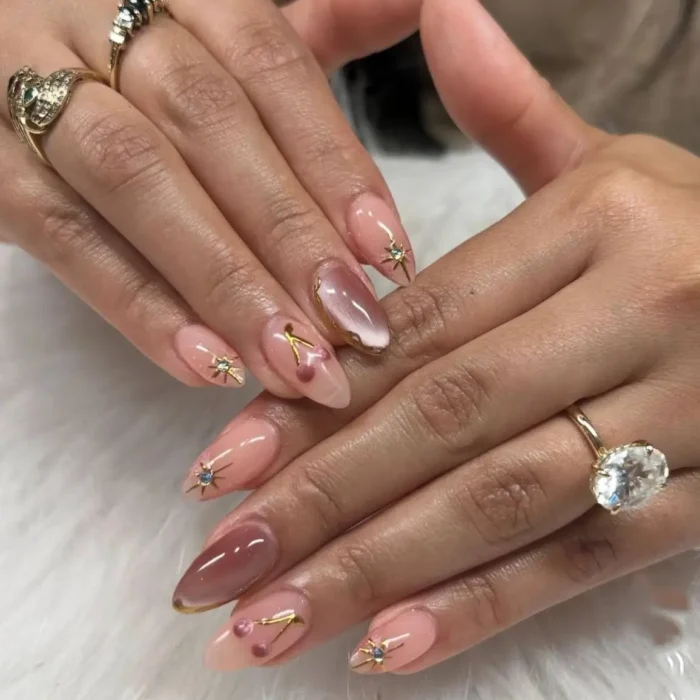 A detailed celestial-themed manicure on long almond nails, featuring pink cat eye effects, gold star charms, and various gems.