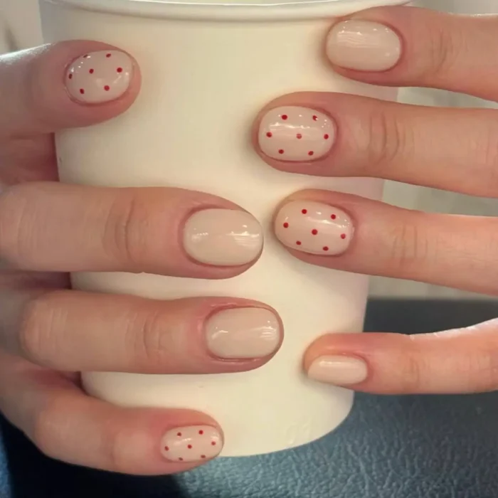 Short nails with a creamy nude gel base, accented by small, evenly spaced red polka dots on each nail.