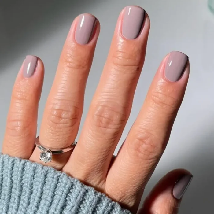 A hand with a flawless manicure in a glossy, muted mauve polish, resting on the cuff of a blue sweater.