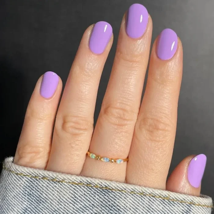 A close-up of a hand with a glossy lilac purple manicure on short, neatly rounded nails against a dark background.