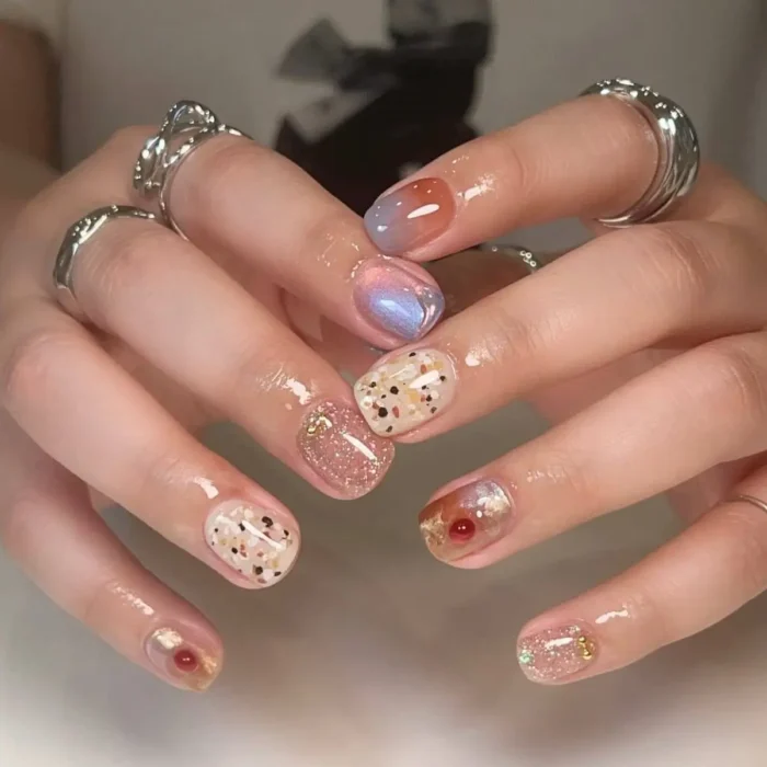 Short nails with a variety of gel designs including soft ombre, glitter with small gold accents, and a speckled pattern in muted tones.