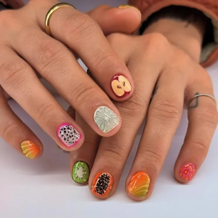 Hands showing a detailed and realistic fruit nail design with a different fruit slice, like apple and dragon fruit, on each nail.