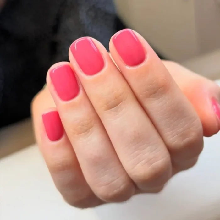A close-up of a hand with a flawless, glossy raspberry-pink manicure on short, neatly filed square nails.