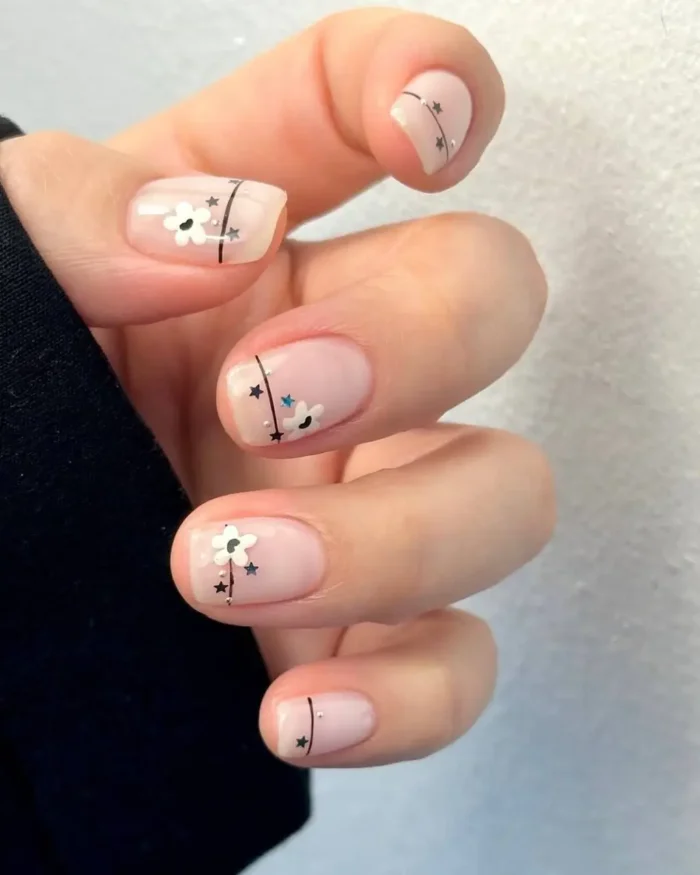 Short, natural-toned nails adorned with small white flowers, thin black lines, and tiny silver star and stud embellishments.