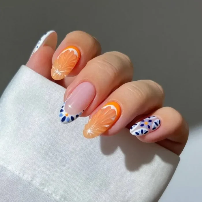 A hand with a chic fruit nail design featuring detailed orange slices and a blue Mediterranean mosaic pattern on almond-shaped nails.