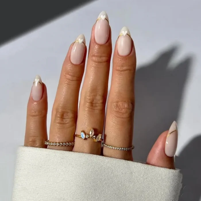A cute white nail design on almond nails, featuring heart-shaped French tips outlined in gold.