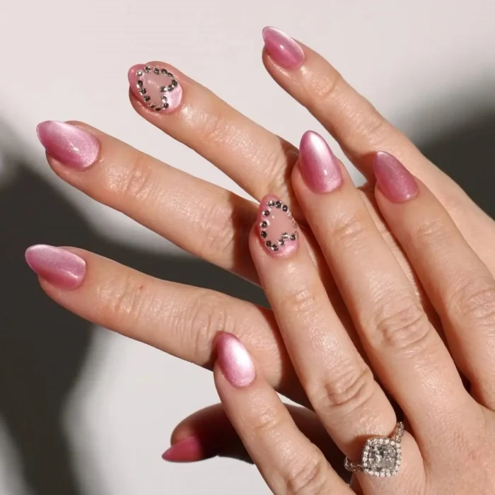 A pink cat eye nail design on almond nails featuring accent nails with crystal hearts on a sheer base.