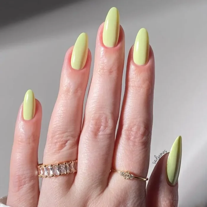 A hand with long, elegant almond nails painted in a glossy, zesty pistachio green polish and adorned with gold rings.