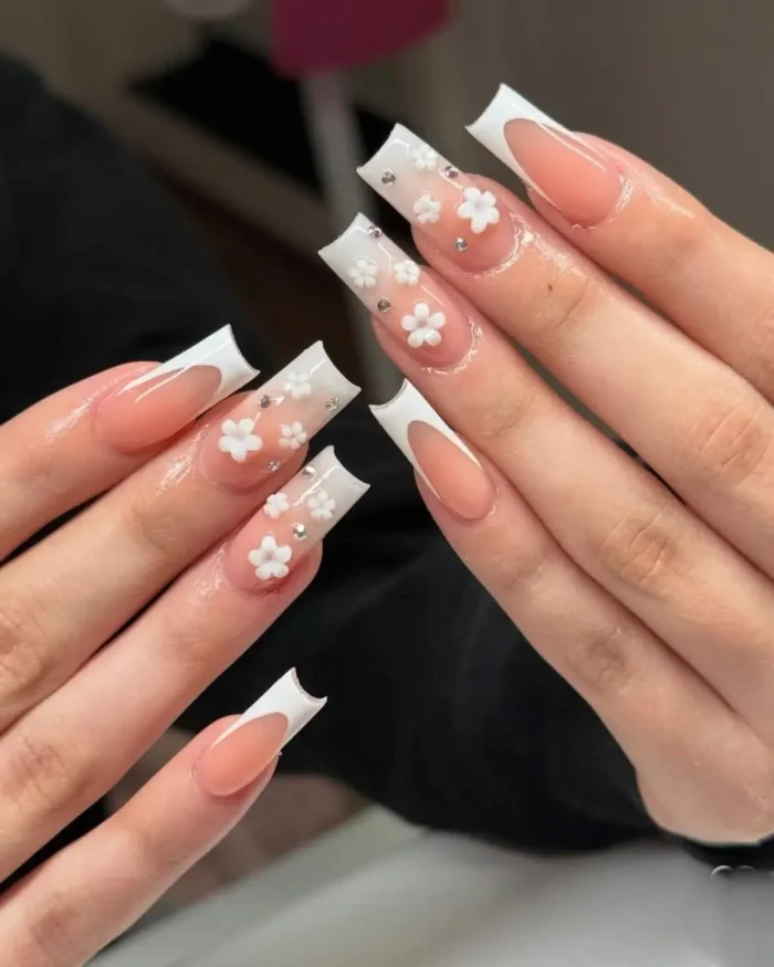 A cute white nail design with 3D white flowers and rhinestones over a French manicure on long square nails.