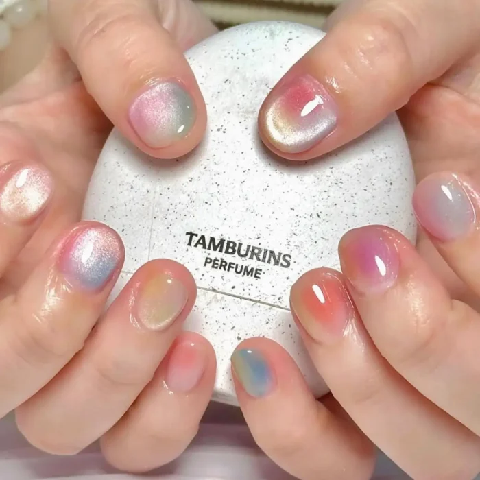 Short, oval-shaped nails with a soft, diffused ombre or 'aura' effect, blending pastel colors like pink, blue, and yellow with a pearlescent finish.