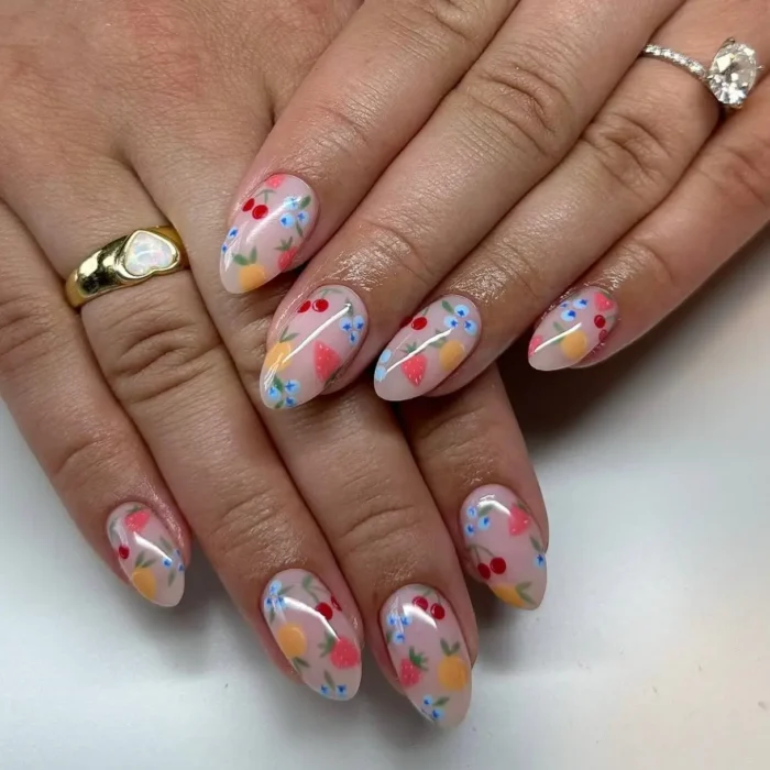 Hands with delicate fruit and wildflower patterned nail art on almond-shaped nails over a sheer pink base.