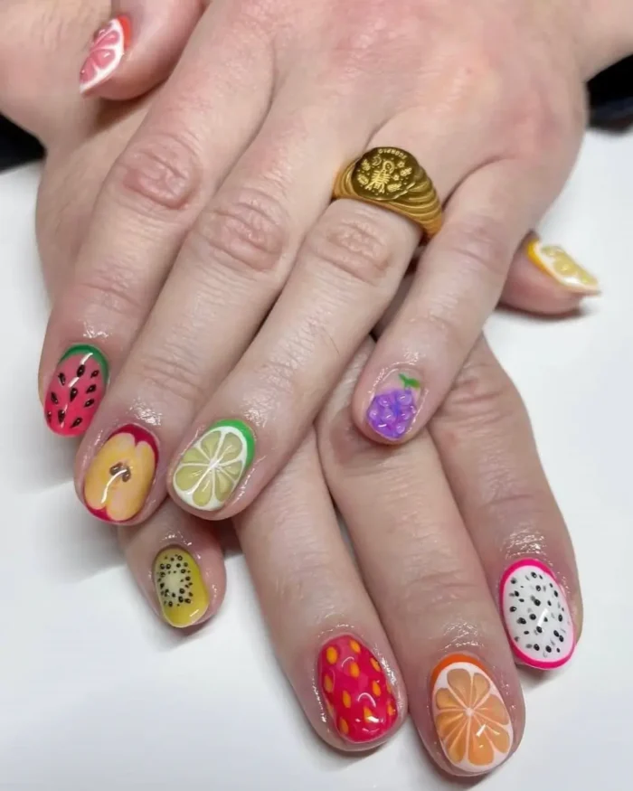 Close-up of hands showing a vibrant, mismatched fruit nail design with detailed art of kiwi, apple, and dragon fruit.