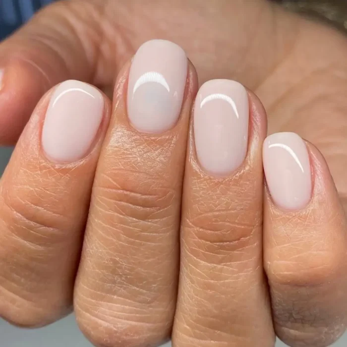 A close-up of a hand showing a glossy, barely-there milky pink manicure on short, neatly shaped nails.