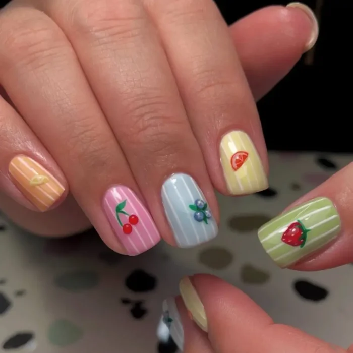 Hands showing a pastel fruit nail design, with each nail featuring a different color pinstripe and a single fruit like a cherry or strawberry.