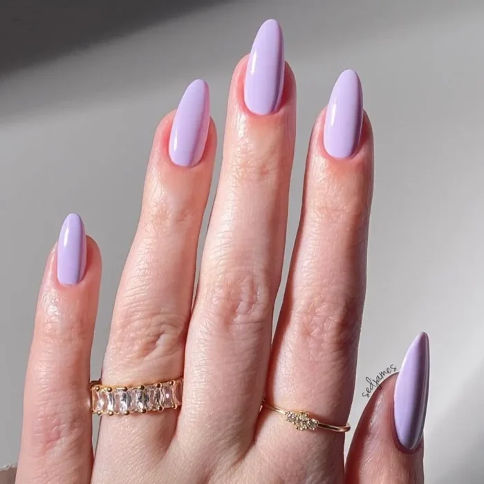 An elegant hand with a glossy lavender cream manicure on long, almond-shaped nails, adorned with delicate gold rings.