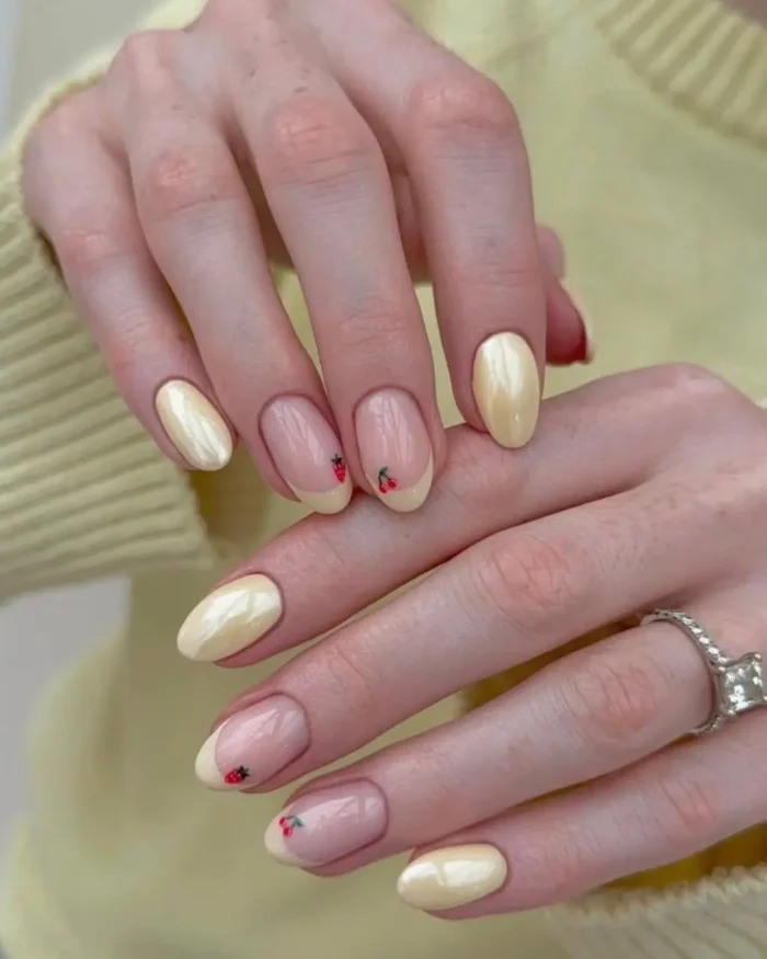 Short, oval nails with alternating designs: some are a creamy, iridescent yellow gel, while others are a sheer nude with hand-painted tiny red strawberries and cherries at the tips.