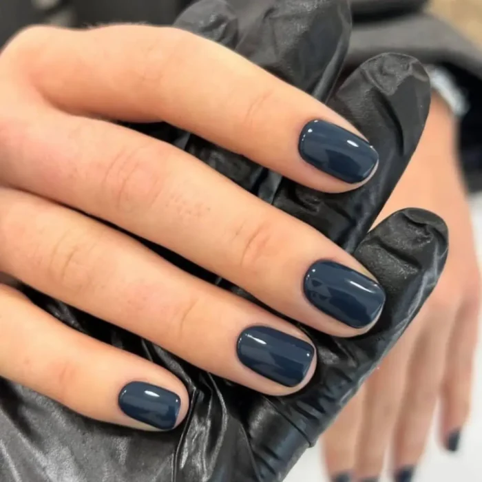 A close-up of a glossy, deep slate-blue manicure on short, square-shaped nails, with the hand resting on a black gloved hand.