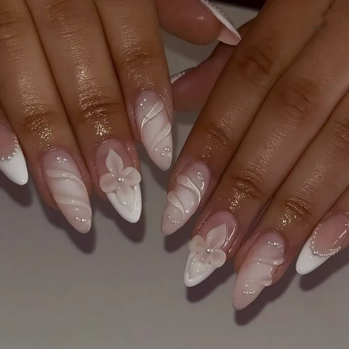 An elegant white nail design on almond nails featuring 3D flowers, sculpted swirls, and pearl details.