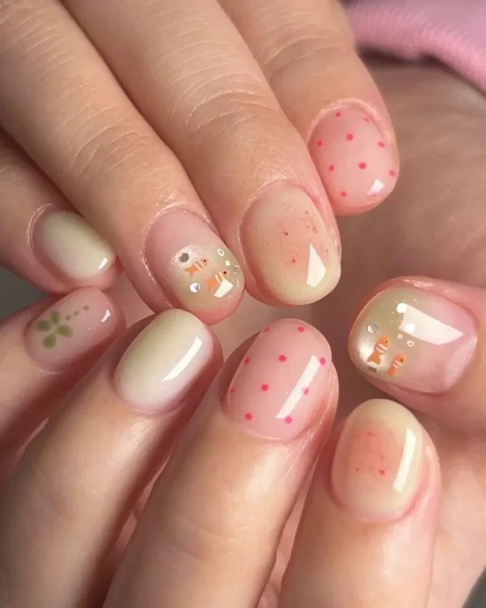 Short, oval nails with a mix of designs: some feature pink polka dots on a nude base, others a light green solid color, and some depict tiny orange goldfish with bubbles on a sheer background.