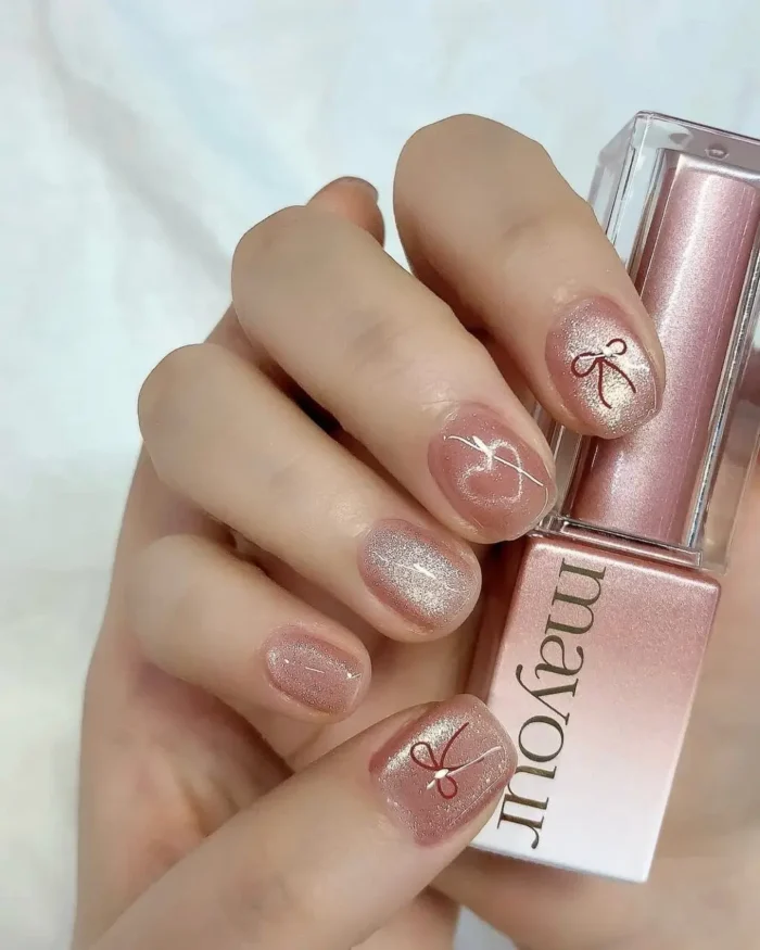 Short nails with shimmering rose gold and iridescent pink gel polish, accented by delicate white line art forming a heart and tiny red bows.