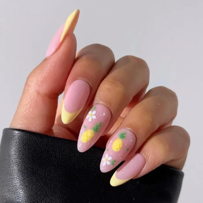 A hand showing a sparkling pink fruit nail design with pineapple and daisy accents and a yellow French tip.
