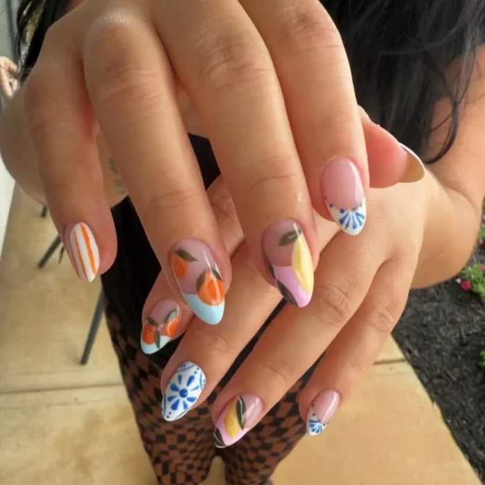 A mismatched fruit nail design showing a mix of orange sprigs, lemon art, and blue tile patterns on almond nails.