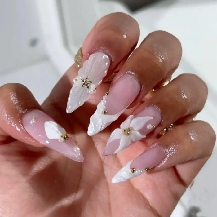 An elegant white nail design on long stiletto nails featuring extravagant 3D sculpted details, pearls, and gold stars.