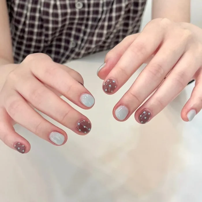 Short, oval nails with alternating designs: some are a solid light blue-grey gel, while others are a warm brown gel with evenly spaced white polka dots.