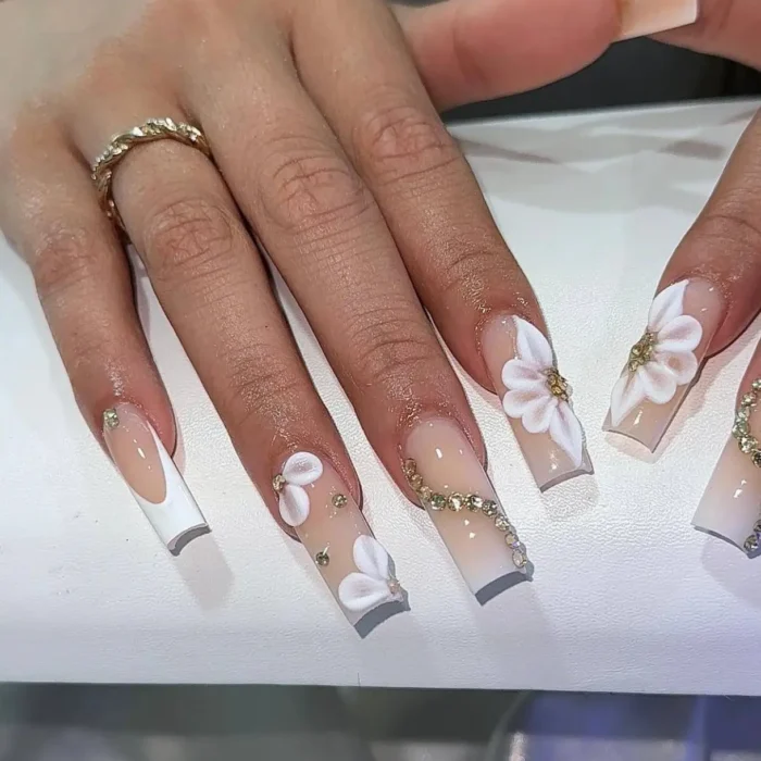 Elegant white nail design on long square nails with 3D flowers, a French tip, and a curving line of gold rhinestones.
