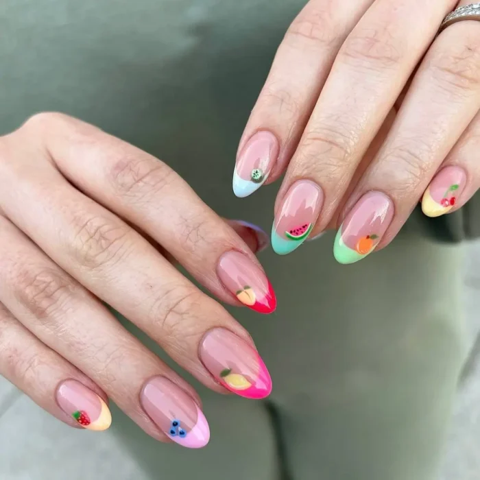 Hands showing a colorful French manicure, with each nail tip painted a different bright color and featuring a tiny fruit design.