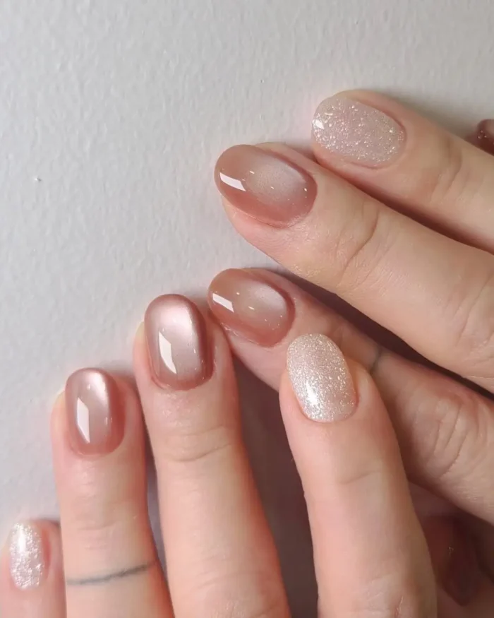 Short nails with a mix of glossy peach-toned gel polish and sparkling glitter on accent nails.