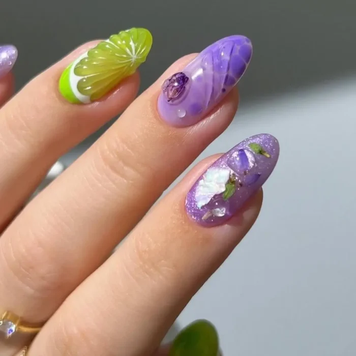 A close-up of a unique manicure with a 3D lime slice accent nail alongside shimmery purple nails with encapsulated flowers.