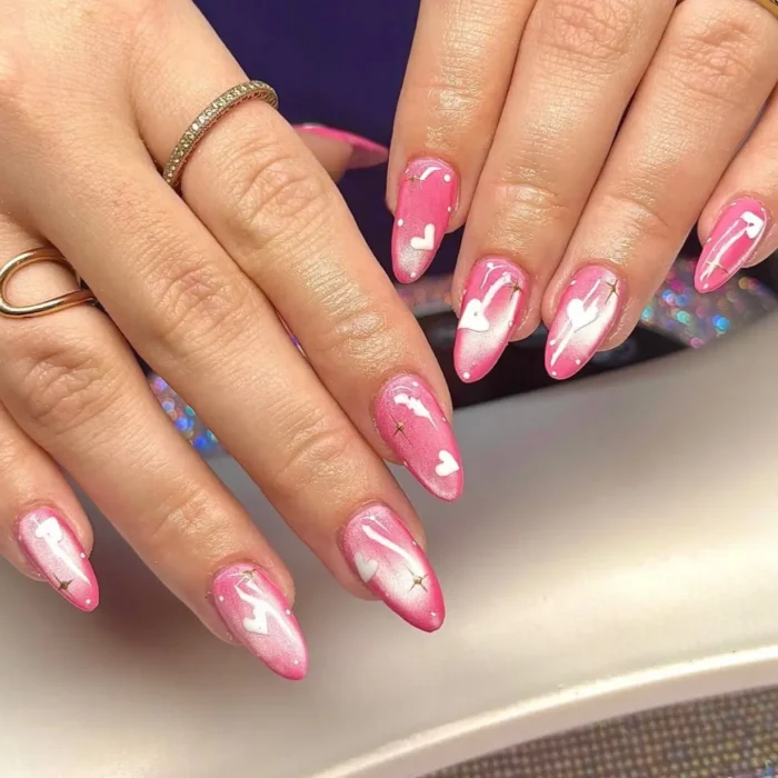 A romantic pink cat eye manicure on long almond nails, featuring hand-painted white hearts and tiny gold star details.