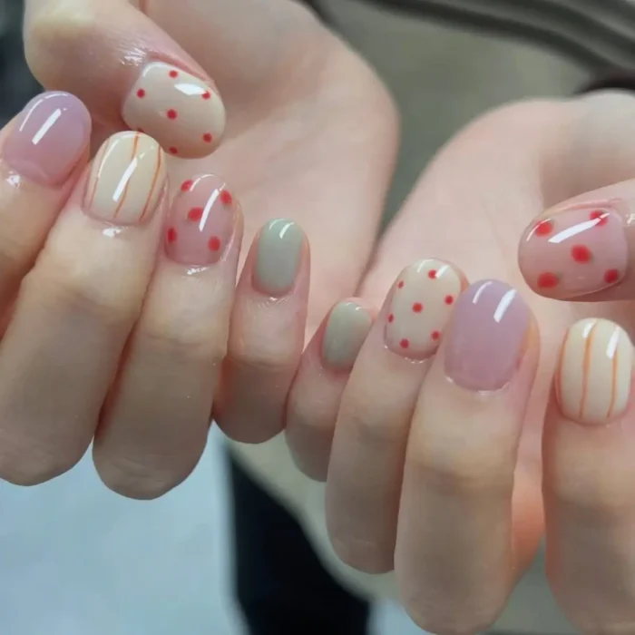 Short, oval nails with a mix of pastel gel designs: solid pink, solid sage green, sheer nude with red polka dots, and sheer nude with thin orange vertical stripes.