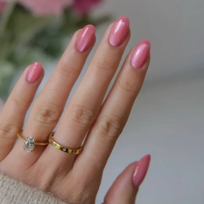 A hand displaying a classic rose-pink cat eye manicure on perfectly shaped oval nails, with gold rings on the fingers.
