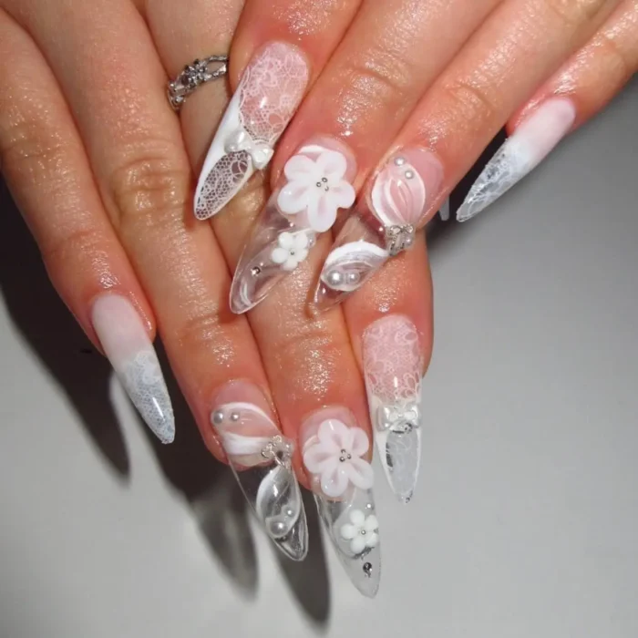 An elegant white nail design on long stiletto nails with intricate 3D flowers, white lace details, and pearls.
