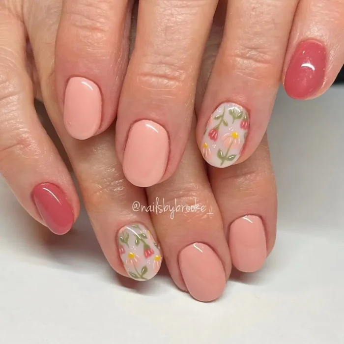 Short, oval nails with a mix of solid peach and muted pink gel polish, and accent nails with delicate hand-painted floral designs featuring small pink and yellow flowers with green stems on a sheer base.