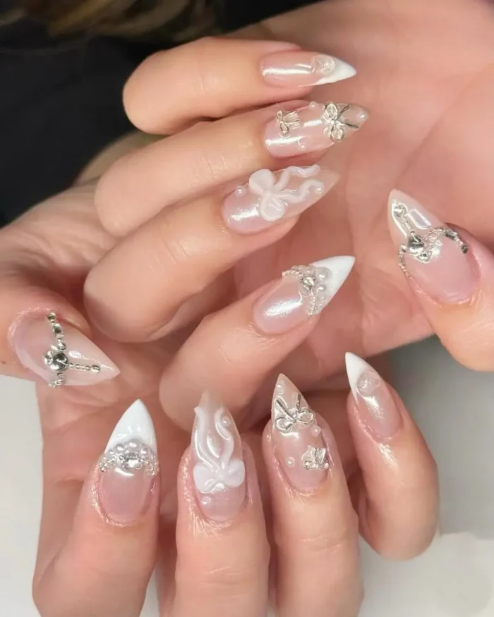 Elegant white nail design on stiletto nails featuring 3D sculpted bows, silver charms, and rhinestone accents.