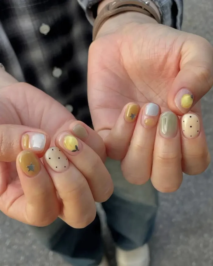 A pair of hands with short nails, each painted with a different whimsical design including pastel colors, fruit, polka dots, and small stars.