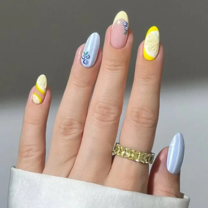 A hand showing a sweet summer fruit nail design on almond nails, featuring lemons, blueberries, and pastel blue stripes.